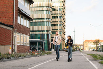 Full length of happy couple riding electric push scooters on street by buildings in city