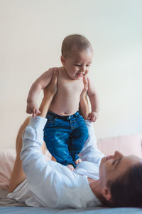 Anonymous mother lying on bed with little baby in blue jeans on stomach between legs on sunny morning