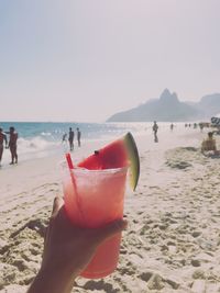 Cropped hand of person holding drink at beach against sky