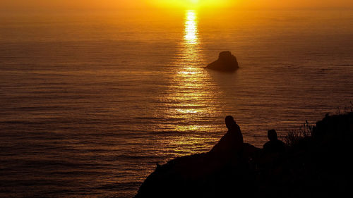 Silhouette person on rock by sea against sky during sunset