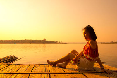 Woman sitting on wooden raft in lake against clear sky during sunset