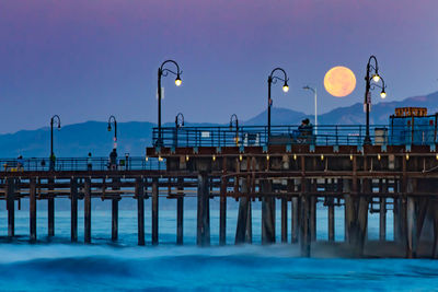 Street lights on pier by sea against sky at dusk