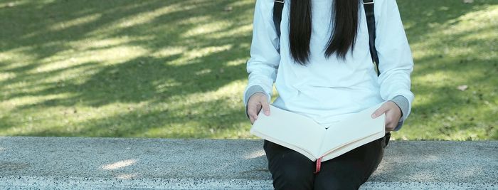 Midsection of woman sitting with book on retaining wall at park