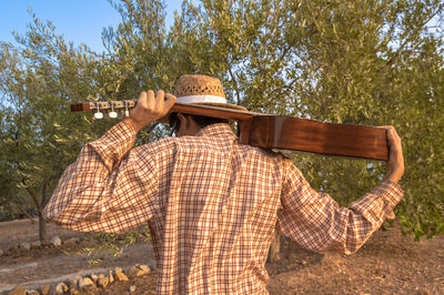 Rear view of man with acoustic guitar standing by trees on field 