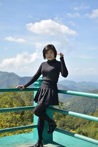 Full length portrait of woman standing against railing and sky