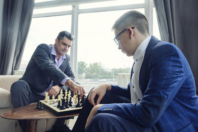 Dad and son are sitting at a table by a wide window and playing chess making moves with pieces