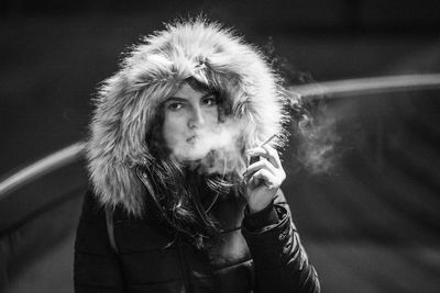 Portrait of young woman warm clothing while smoking cigarette on footpath at night