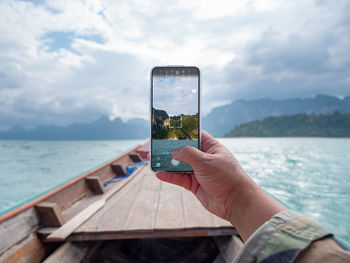 Hand holding smart phone by sea against sky