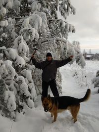 Portrait of smiling man with axe standing by dog and trees on snow during winter
