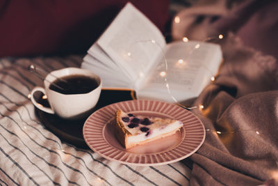 Cup of black tea, tasty fruit cake and open book with glowing lights in bed closeup. 