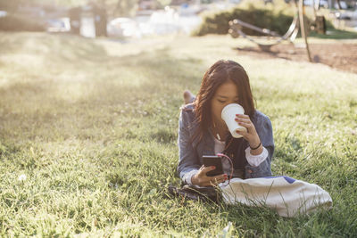 Teenage girl drinking coffee while using smart phone on grass