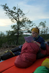 Man using mobile phone while sitting by tree against sky
