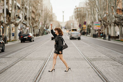 Side view of successful businesswoman in retro leather suit waving hand and trying to catch taxi while crossing tram rails on city street