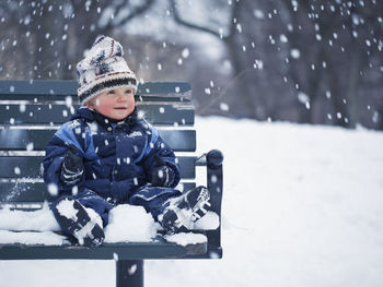 Baby boy sitting on bench and looking at falling snow