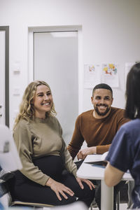 Happy pregnant woman discussing with female doctor while sitting by man in hospital