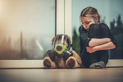 Girl with teddy bear wearing mask sitting against window