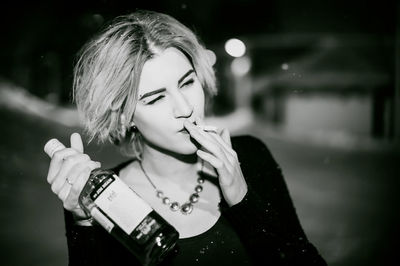 Close-up of young woman smoking cigarette while holding alcohol bottle at night