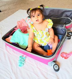 High angle view of cute baby girl sitting in suitcase on floor at home