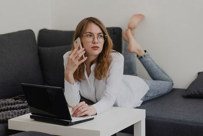 Attractive young woman working from home - female entrepreneur sitting on sofa with laptop computer and checking cell phone from comfort of home