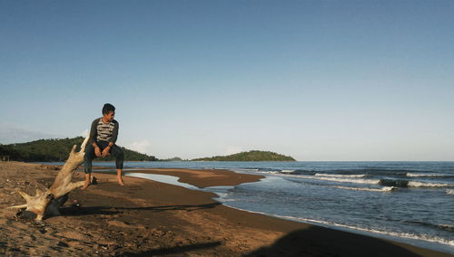 Man sitting on driftwood at beach against sky