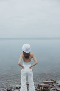 Woman looking out at sea wearing a white bucket hat, white flared jeans and a blu bathing suit. 