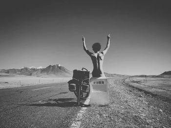 Naked man with obscene gesture sitting on sign by bicycle amidst road against clear sky