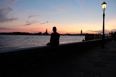 Silhouette woman sitting by sea against sky during sunset