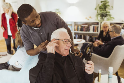 Male healthcare worker talking with senior man while cutting hair in retirement home