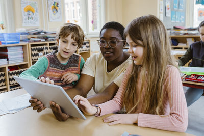 Female teacher sitting amidst schoolboy and schoolgirl using digital tablet sitting at desk in classroom