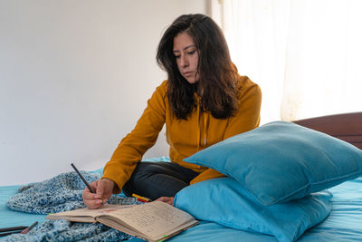 Young woman writing in book while sitting on bed at home