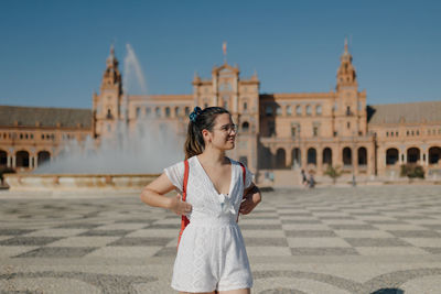 Young tourist woman wearing a white dress is looking away and smiling while standing in seville.