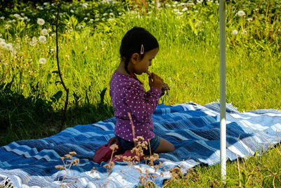 Side view of girl eating while kneeling on picnic blanket amidst grass