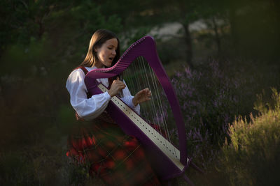 Beautiful woman playing celtic harp while sitting on grassy field