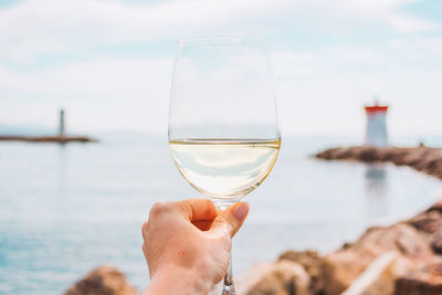 Woman hand with white wine glass on a sea, stones and lighthouse background. nice, french riviera