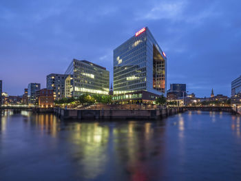 Illuminated buildings by river against sky in city at dusk