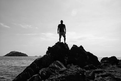 Rear view of man standing on rock at sea shore against sky