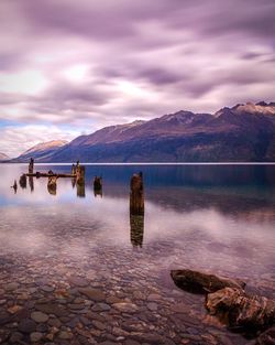 Wooden posts in river by rocky mountains against cloudy sky