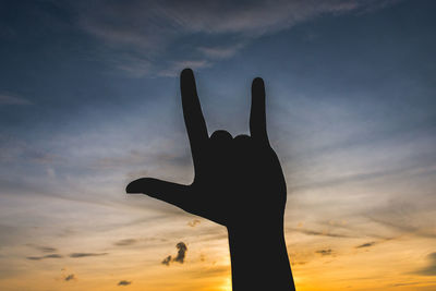 Cropped image of hand gesturing horn sign against sky during sunset