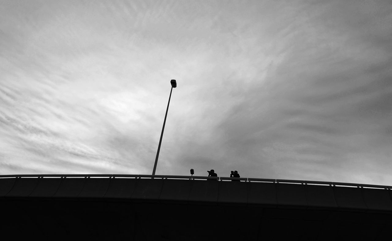 low angle view, sky, architecture, built structure, silhouette, cloud - sky, street light, men, cloudy, railing, cloud, building exterior, connection, lifestyles, leisure activity, outdoors, unrecognizable person, bridge - man made structure