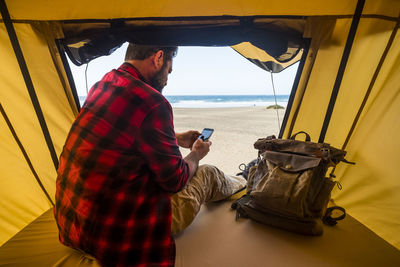 Rear view of man using smart phone sitting in tent against sea