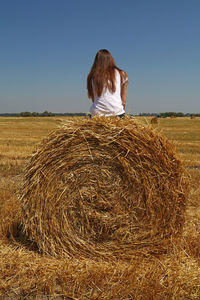 Rear view of woman sitting on hay bale at farm against clear blue sky