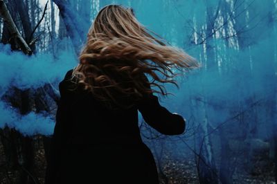 Rear view of woman with tousled hair against blue distress flare in forest