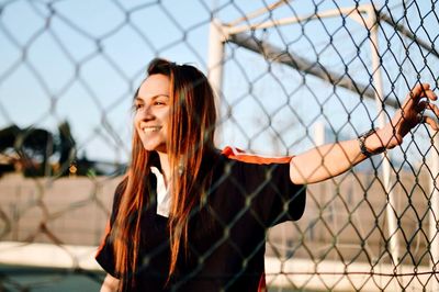 Portrait of smiling young woman standing by chainlink fence
