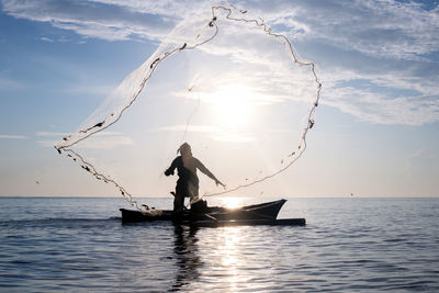 Silhouette fisherman throwing fishing net in sea against sky during sunset