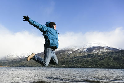 Excited hiker jumping by river against sky