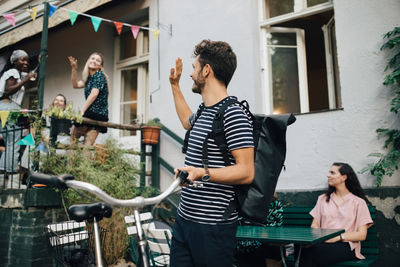 Smiling young man waving hand while standing with bicycle in backyard