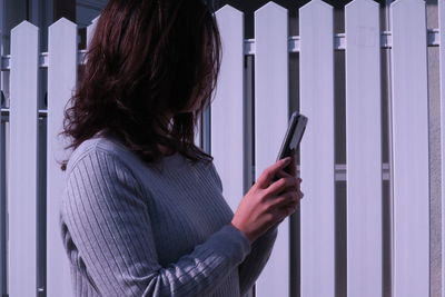 Side view of woman using mobile phone while standing by fence