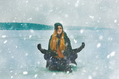 Full length of young woman enjoying snowfall while sitting on snowfield