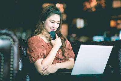 Young woman using mobile phone while sitting on laptop