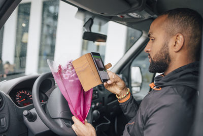 Male messenger with bouquet of flowers looking at smart phone while sitting in delivery van